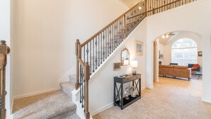 Un escalier avec une rampe en bois et des balustres noires mène au deuxième étage. Une console ornée d’objets décoratifs est placée à côté de l’escalier.