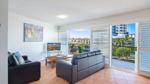 Un salon moderne avec un canapé, une table basse et un téléviseur posé sur un meuble. Il y a un balcon d’où l’on peut admirer les bâtiments et les palmiers.