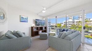 A modern living room with grey sofas, a wooden bookshelf, a flat-screen TV, and a balcony with a view of buildings and greenery. - 2 Bedroom Apartment Sea View Second Floor (Sunshine Coast)