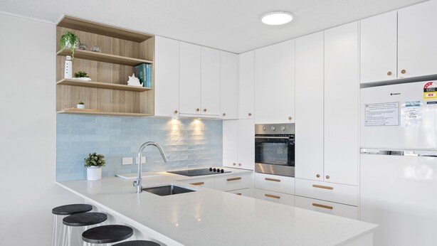 A modern kitchen with white cabinetry, a central island with stools, a stainless steel sink, and a built-in oven. - 2 Bedroom Apartment Sea View Second Floor (Sunshine Coast)