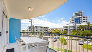 A balcony with a white table and chairs, overlooking a street with cars and buildings. - 2 Bedroom Apartment Sea View Second Floor (Sunshine Coast)