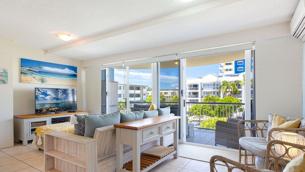 A modern living room with a TV, sofa, and dining area, featuring large windows and a balcony with a view of buildings and greenery.