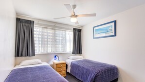 Une chambre d’hôtel avec deux lits simples, une table de chevet avec une lampe, un ventilateur de plafond, une fenêtre avec des rideaux et un tableau encadré au mur.