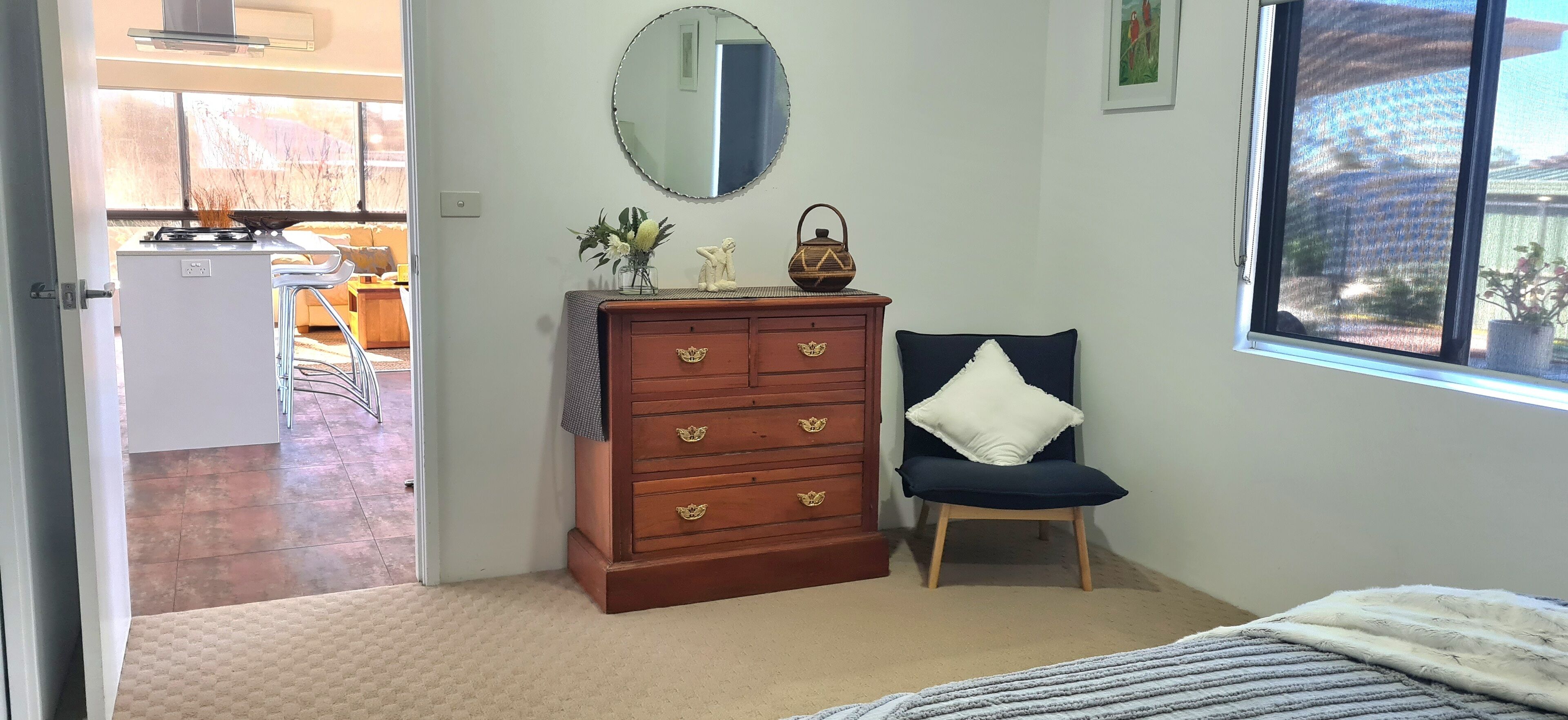 A bedroom with a wooden dresser, a chair with a pillow, a bed with a blanket, a mirror, and a window with a view.