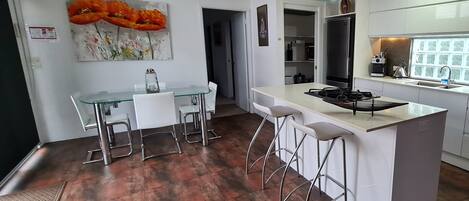Cozinha moderna com ilha central, banquetas e área de jantar com mesa de vidro e cadeiras.