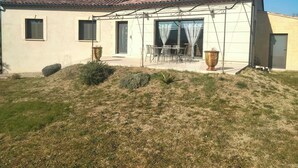 Une maison de plain-pied avec un toit en tuiles, une terrasse avec des meubles d’extérieur et une grande fenêtre avec des rideaux.