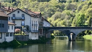 Un pont de pierre enjambe une rivière paisible, avec des bâtiments d’un côté et une colline boisée de l’autre.