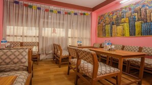 A dining area with wooden furniture, a large painting on the wall, and a window with curtains. - MeroStay 211 Hotel DN (Kathmandu)