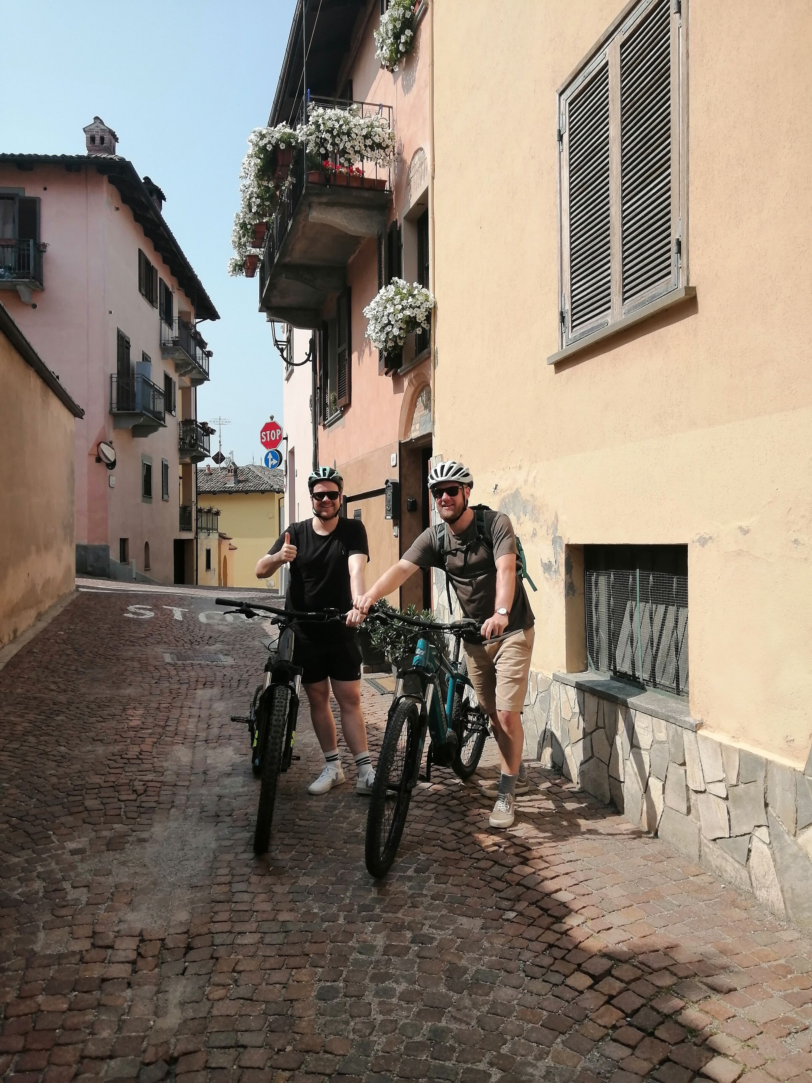 Deux personnes à vélo dans une rue étroite, pavée de pierres, bordée d  ’immeubles avec des balcons, et un panneau de signalisation indiquant un arrêt.