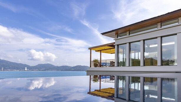 A modern building with a glass facade, a wooden deck, and a view of the sea. - Villa Fah Sai (Kamala)