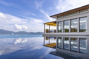 A modern building with a glass facade, a wooden deck, and a view of the sea. - Villa Fah Sai (Kamala)