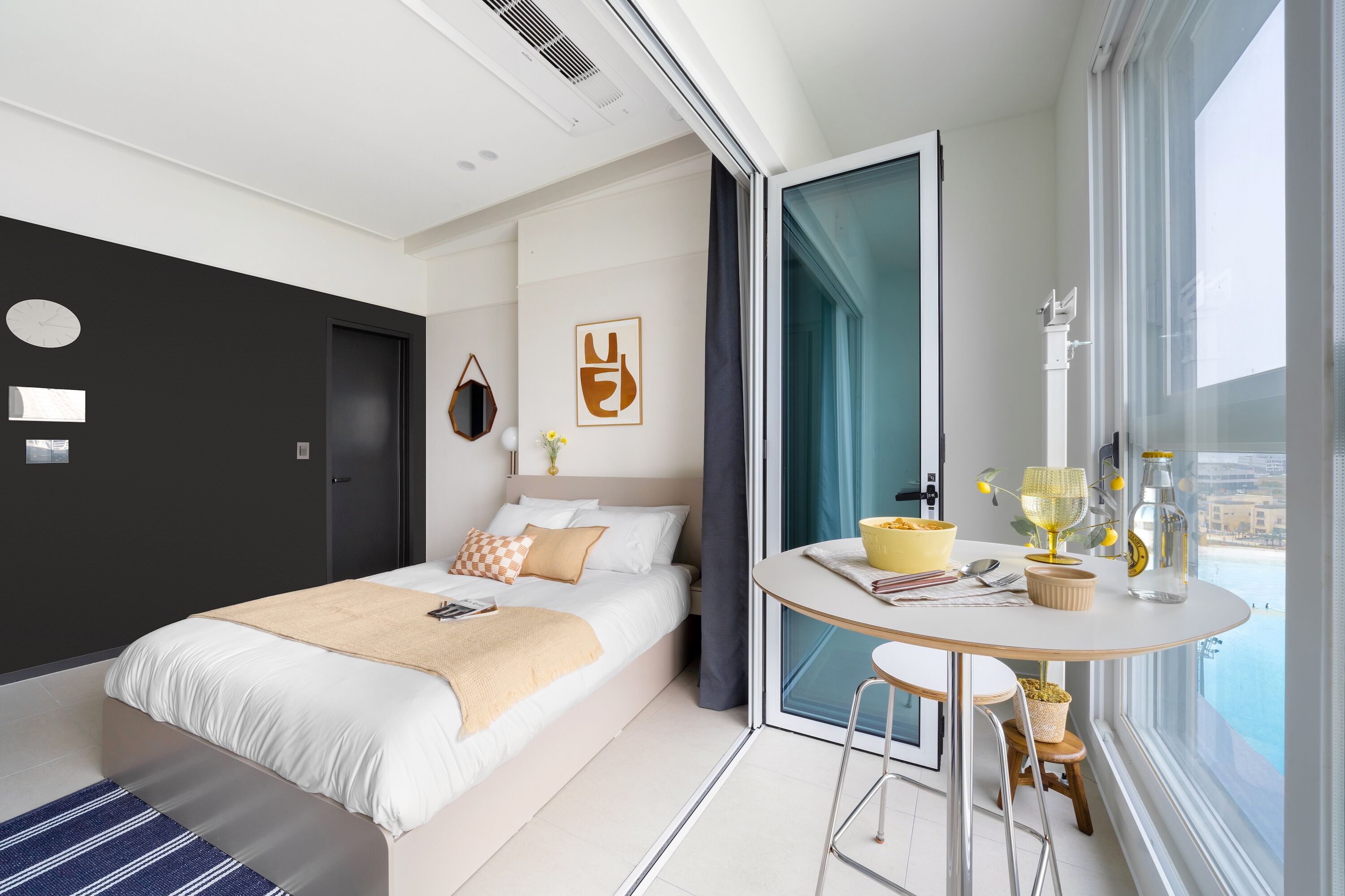 A modern hotel room with a bed, a round dining table with chairs, a window with a view, and a door.