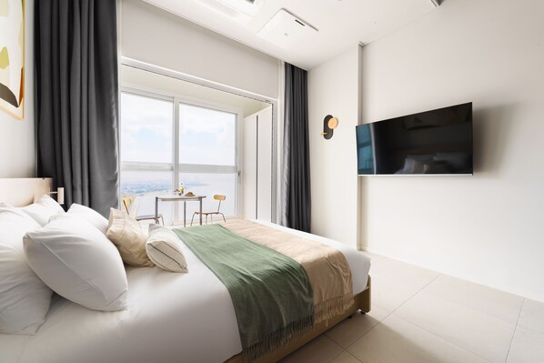 A modern hotel room with a large bed, a flat-screen TV mounted on the wall, a window with a view, and a small table with chairs.
