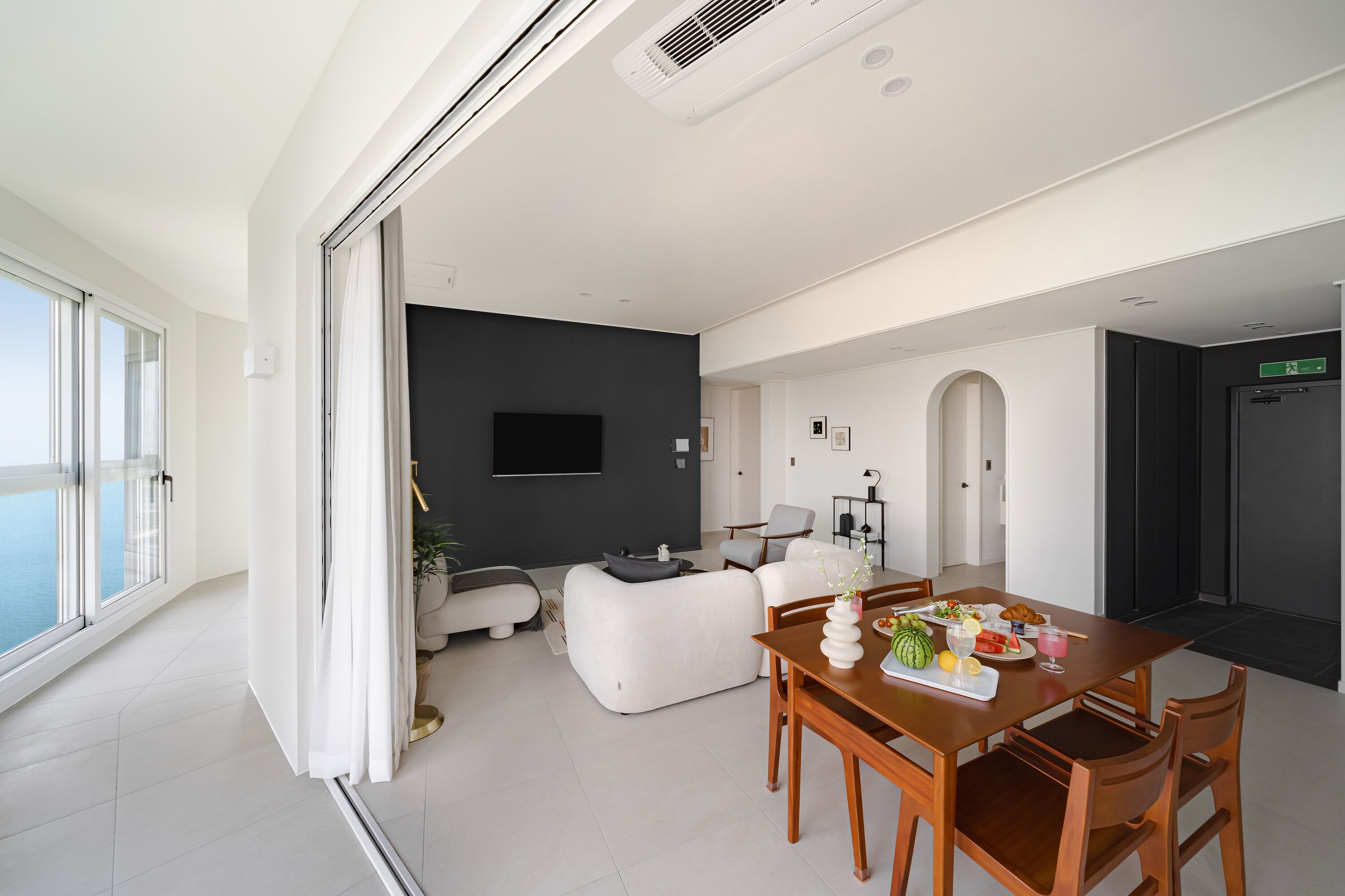 A modern living room with a dining area, a flat-screen TV, and a large window overlooking a body of water.