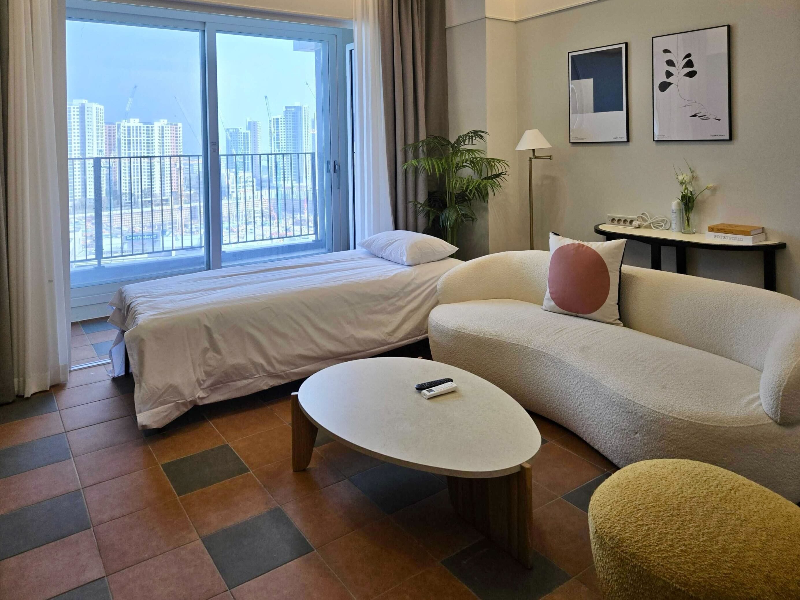 A hotel room with a bed, a sofa, a small table, and a view of the cityscape through a large window.