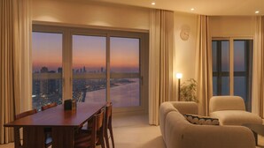 Une chambre d’hôtel moderne dot ée d’une grande baie vitrée panoramique, d’un coin repas et d’un espace salon offrant une vue sur le paysage urbain au coucher du soleil.