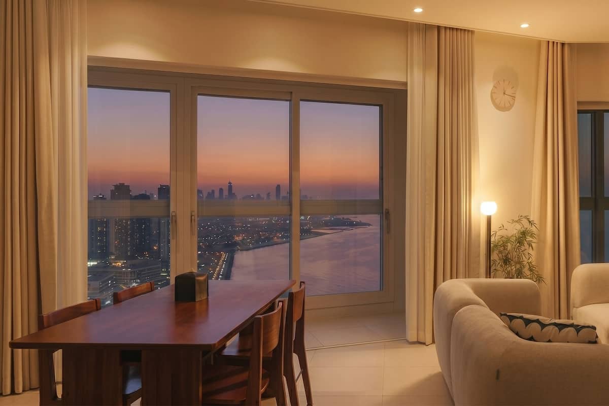 Habitació d'hotel moderna amb un gran finestral panoràmic, zona de dinar i espai d'estar amb vista a un paisatge urbà al capvespre.