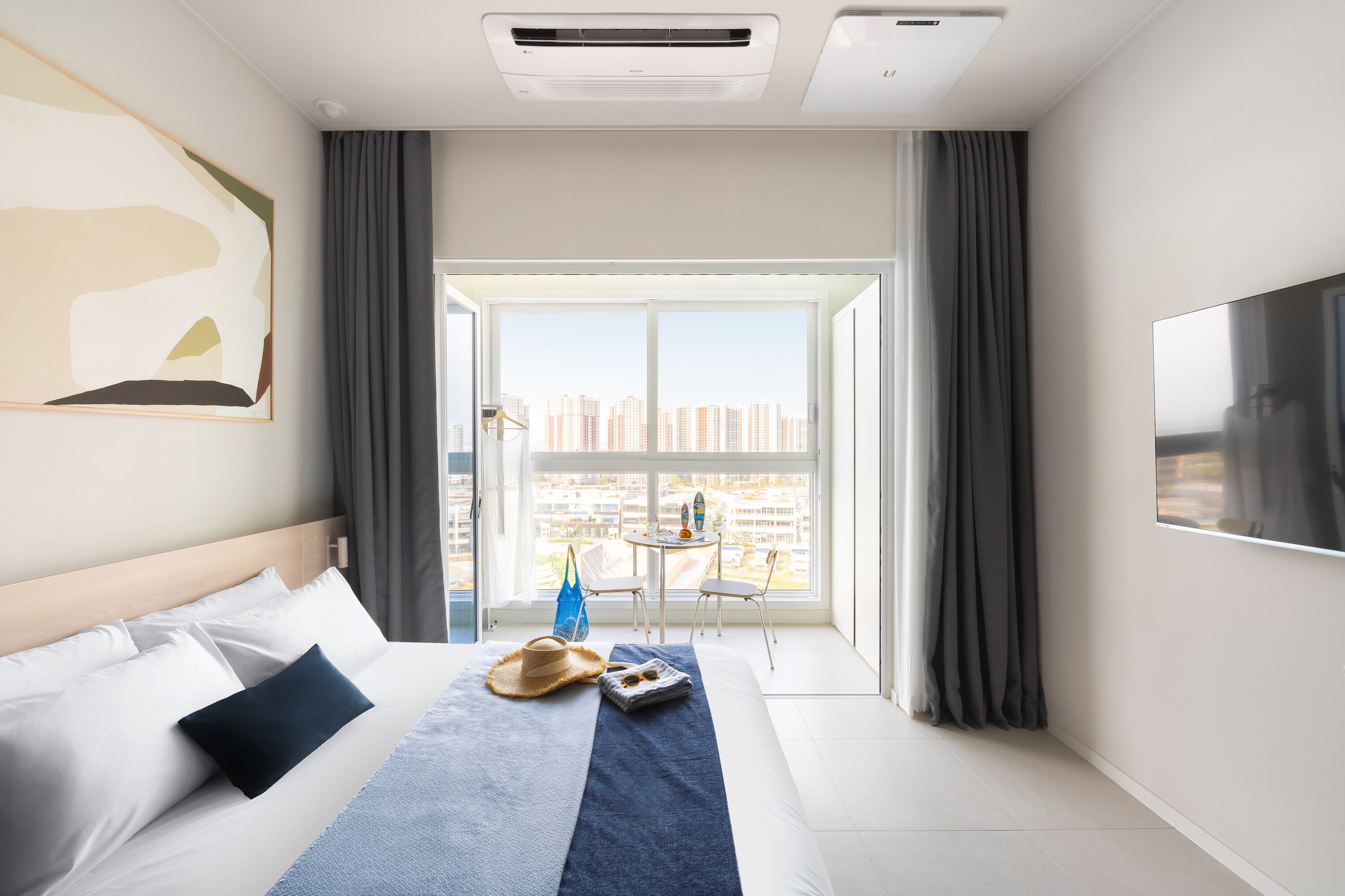 Quarto de hotel com cama, janela ampla, televisão e vista para a paisagem urbana.