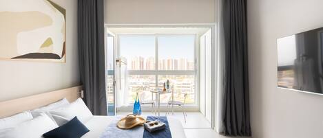 Quarto de hotel com cama, janela ampla, televisão e vista para a paisagem urbana.