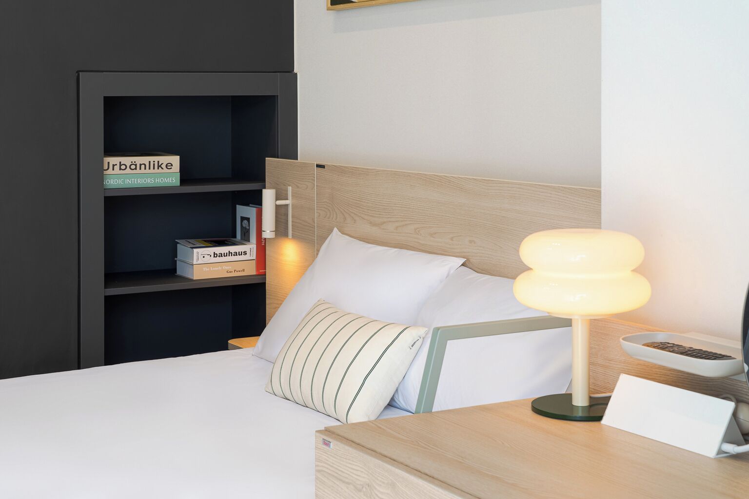 A neatly arranged hotel room with a bed, a bedside table with a lamp, a shelf with books, and a sign indicating the brand 'Urbanike'.