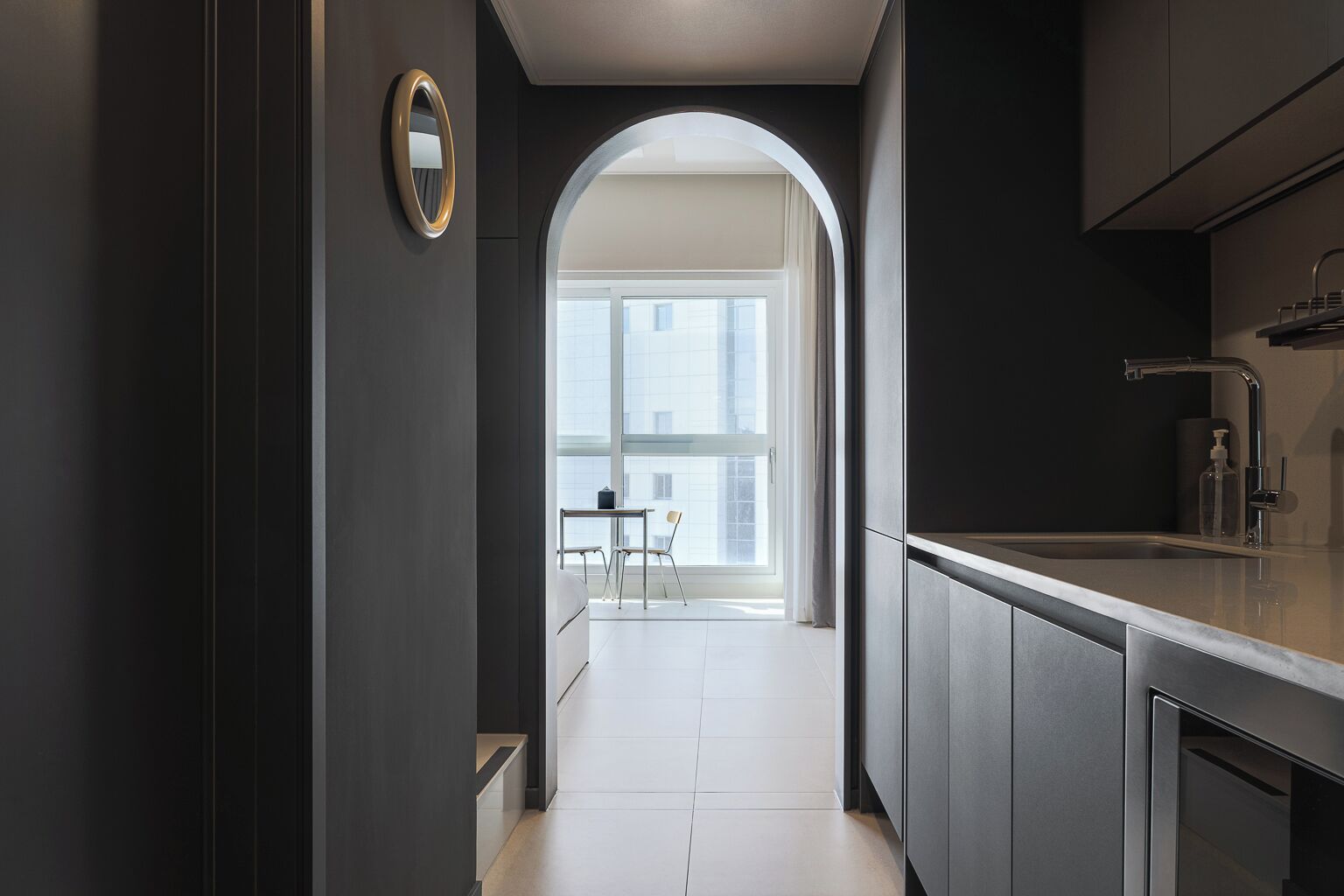 A modern kitchen with dark cabinetry, a stainless steel sink, and a view through an archway to a balcony with a table and chairs.