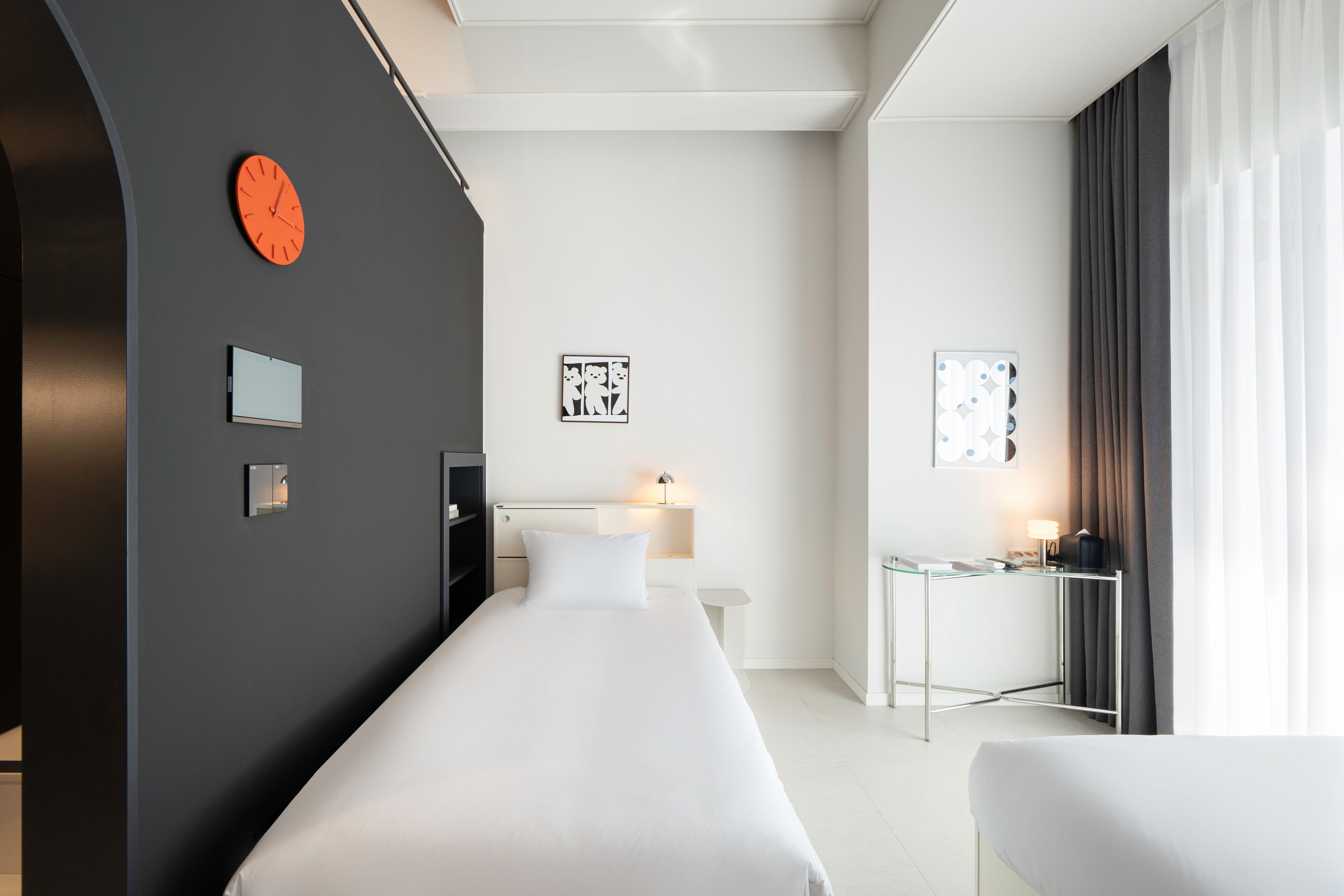 Ein modernes Hotelzimmer mit einem großen Bett, einem Schreibtisch und einem Stuhl. An der Wand hängt eine Uhr und ein kleiner Tisch mit einer Lampe steht bereit.