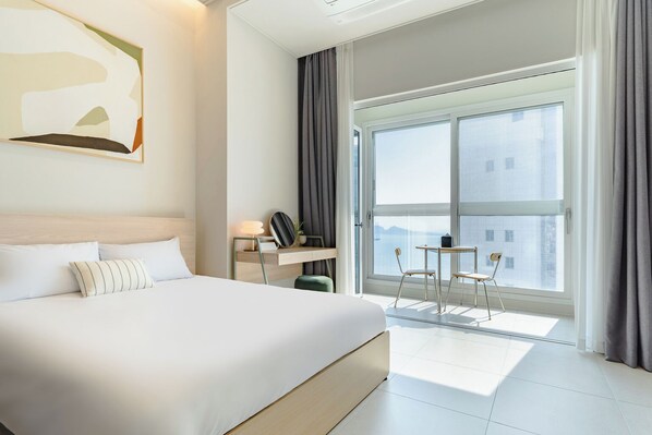 Een moderne hotelkamer met een groot bed, een bureau met stoel, een klein tafeltje met twee stoelen en een groot raam met uitzicht op de stad.