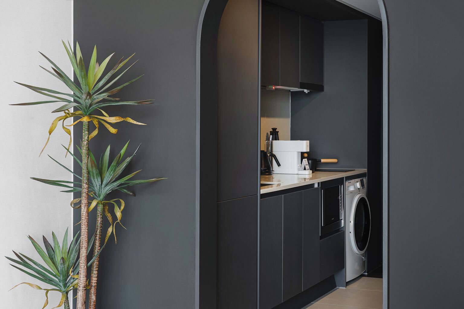 A modern kitchen with dark cabinetry, a washing machine, and a coffee machine.