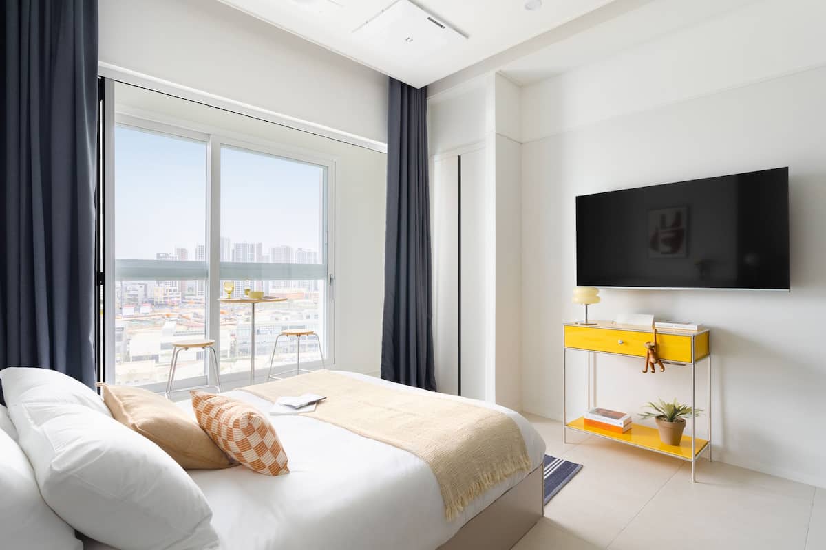 Habitació d'hotel moderna amb un llit gran, una televisió penjada a la paret, una taula petita amb una cadira i vista a la ciutat per la finestra.