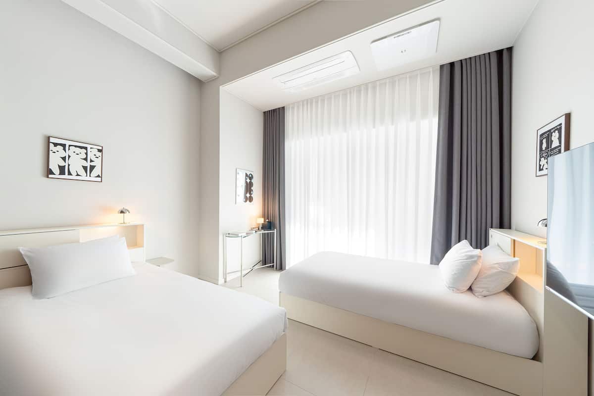 Una habitació d'hotel moderna amb dos llits, una taula de treball i una finestra gran amb cortines.