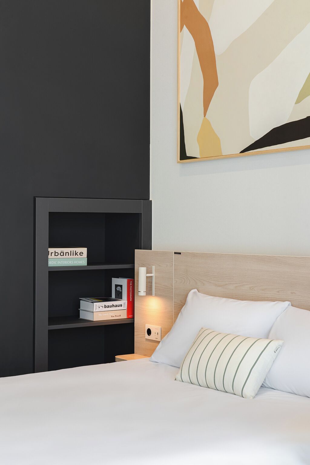 A modern hotel room with a dark wall, a wooden headboard, a white bed with striped pillows, a bedside table with a lamp, and a bookshelf with books.