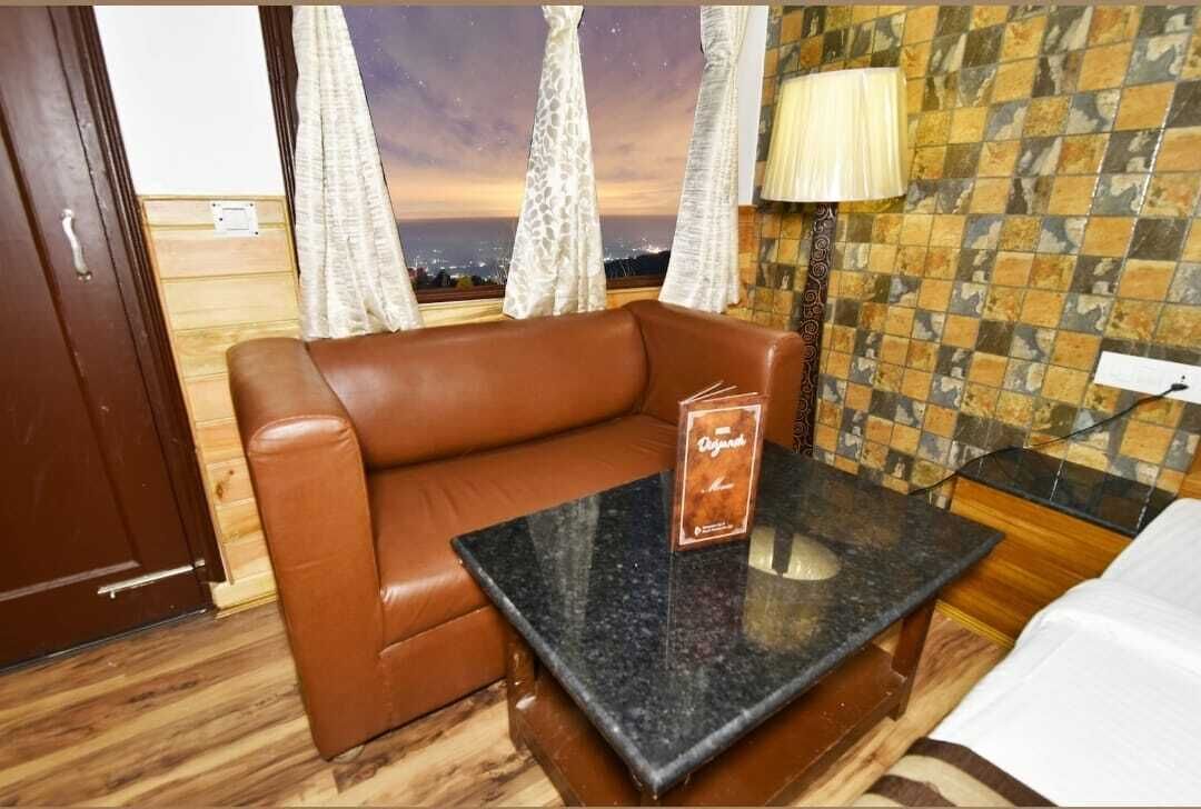 Ein Zimmer mit einem braunen Ledersofa, einem kleinen Tisch mit einem Schild, einer Lampe und Blick auf die Stadt im Abendrot durch Vorhänge.