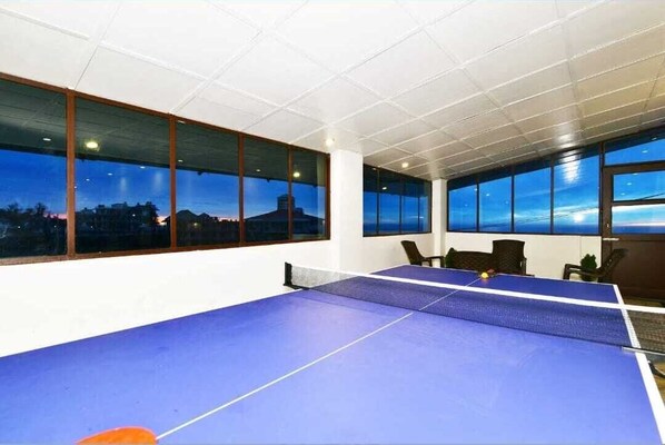 A ping pong table in a room with large windows offering a view of a cityscape at dusk.