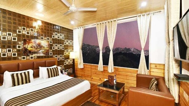 A hotel room with a bed, a sofa, a small table, a ceiling fan, a large window with curtains, and a wall decorated with picture frames. - Hotel Divyansh By VRB Hotels (Dharamshala)