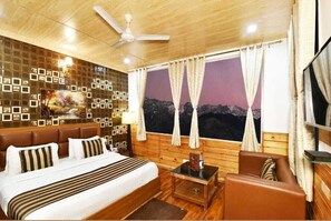 A hotel room with a bed, a sofa, a small table, a ceiling fan, a large window with curtains, and a wall decorated with picture frames.