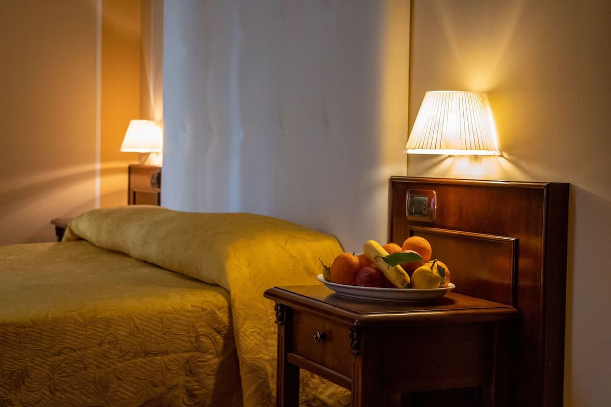 A hotel room with a bed, a bedside table with a lamp, and a bowl of fruit.