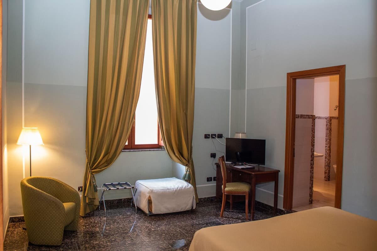 A hotel room with a bed, a desk with a television, a chair, a lamp, and a window with curtains.