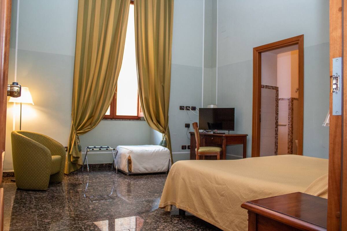 A hotel room with a bed, a desk with a chair, a small table, a lamp, and a television.