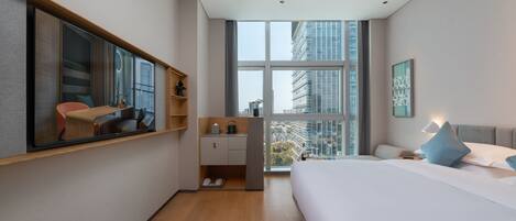 Habitación de hotel moderna con una cama grande, un escritorio con silla, una kitchenette pequeña y un ventanal con vistas a la ciudad.