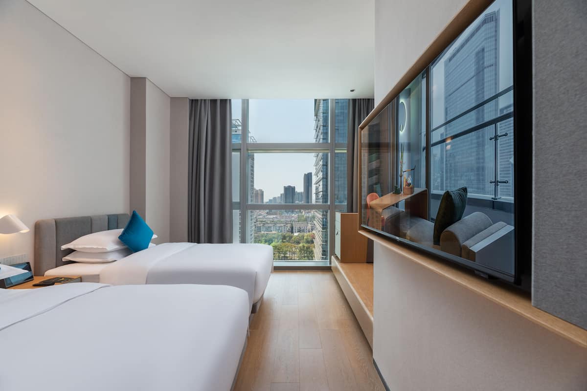 Una habitació d'hotel amb dos llits, una finestra gran que dona a una vista de la ciutat, un televisor i una taula petita amb una làmpada.