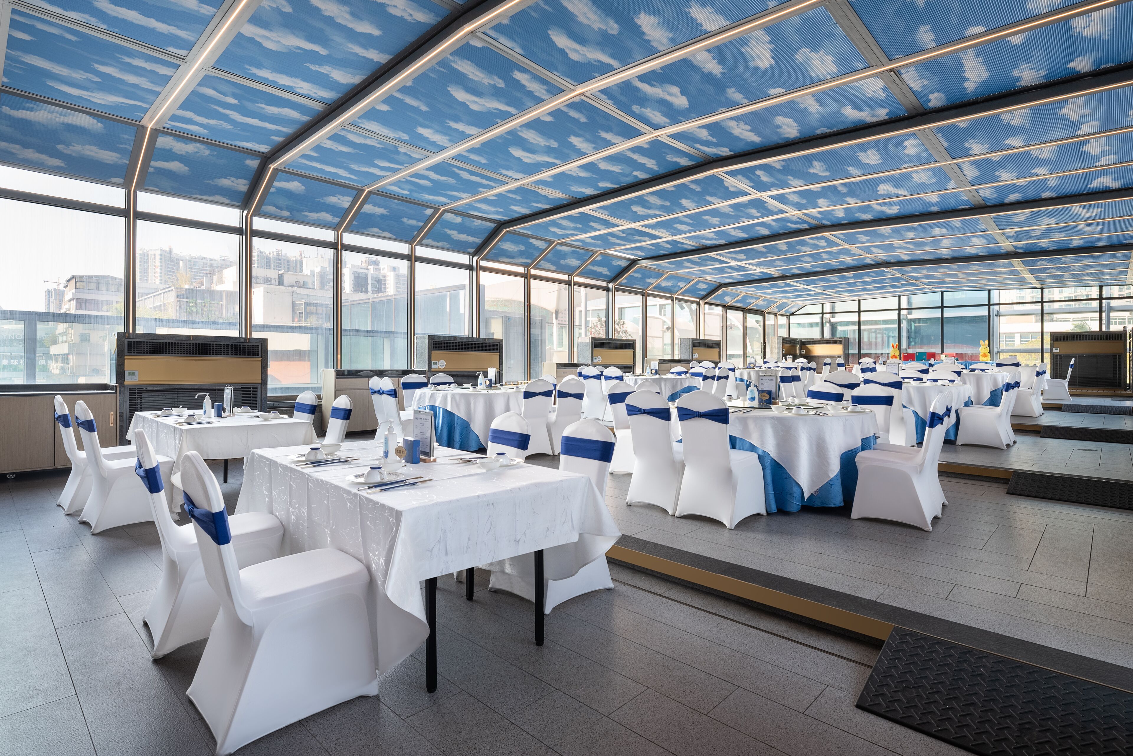 A spacious indoor event space with tables and chairs, large windows, and a high ceiling with a patterned design.