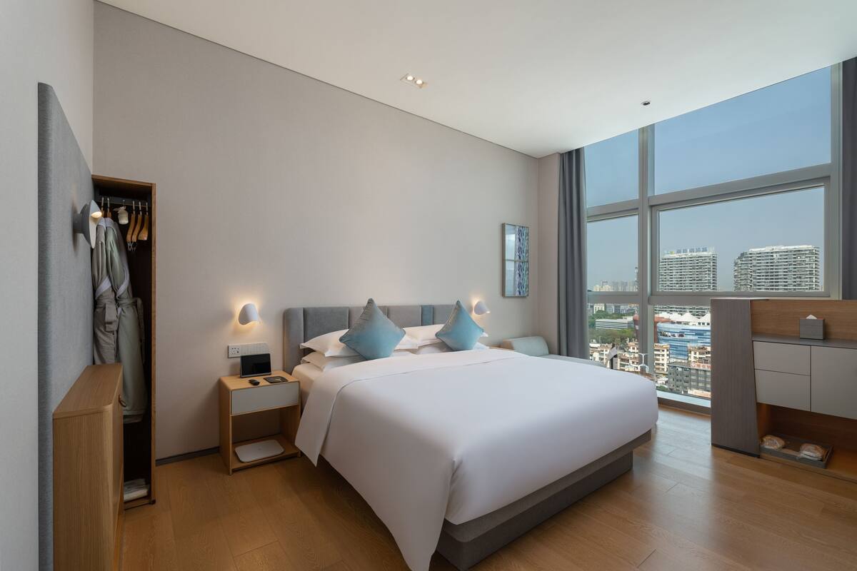 Une chambre d’hôtel moderne dotée d’un grand lit, de tables de chevet, d’une armoire et offrant une vue sur la ville grâce à de grandes fenêtres.