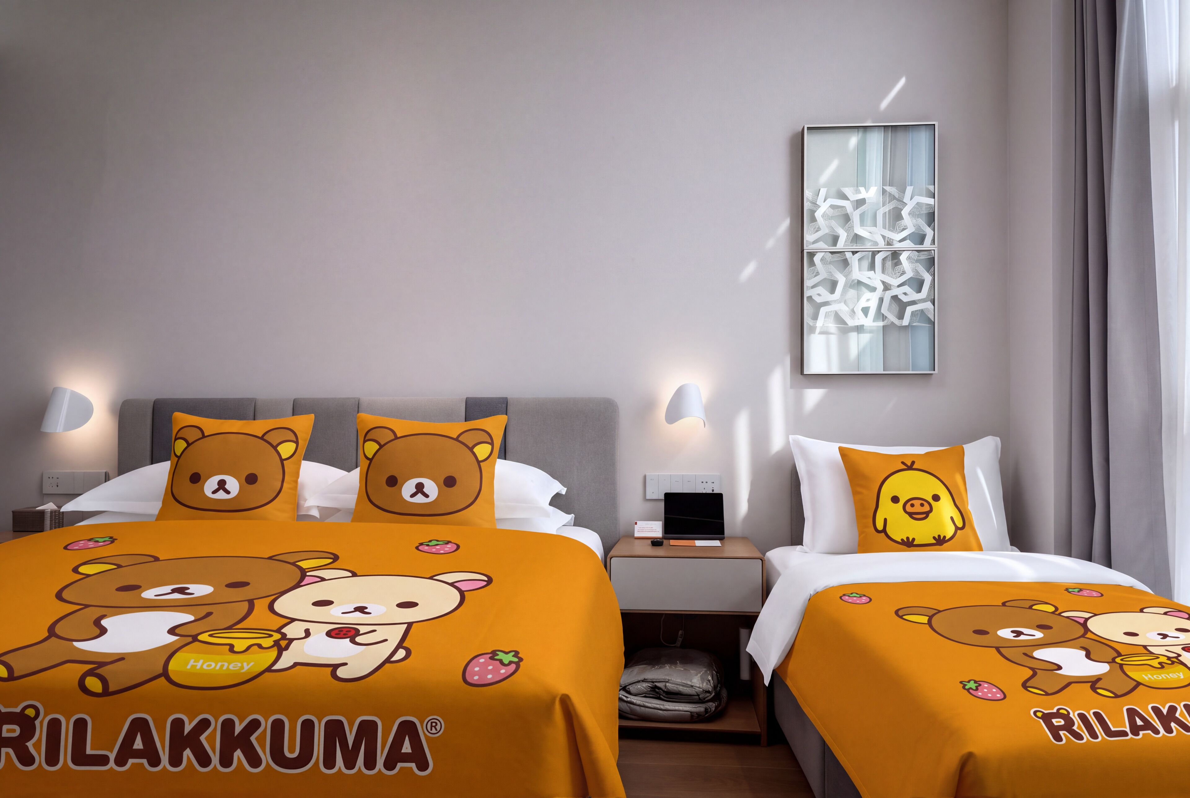 Une chambre d’hôtel avec deux lits, chacun recouvert d’une couette à motifs d’ours. On y trouve une table de chevet avec une lampe, un luminaire fixé au mur et une fenêtre avec des rideaux.