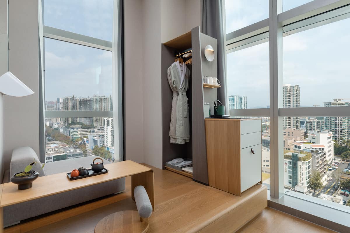 Une chambre d’hôtel moderne avec une grande fenêtre, un bureau en bois, une robe suspendue et un petit placard.