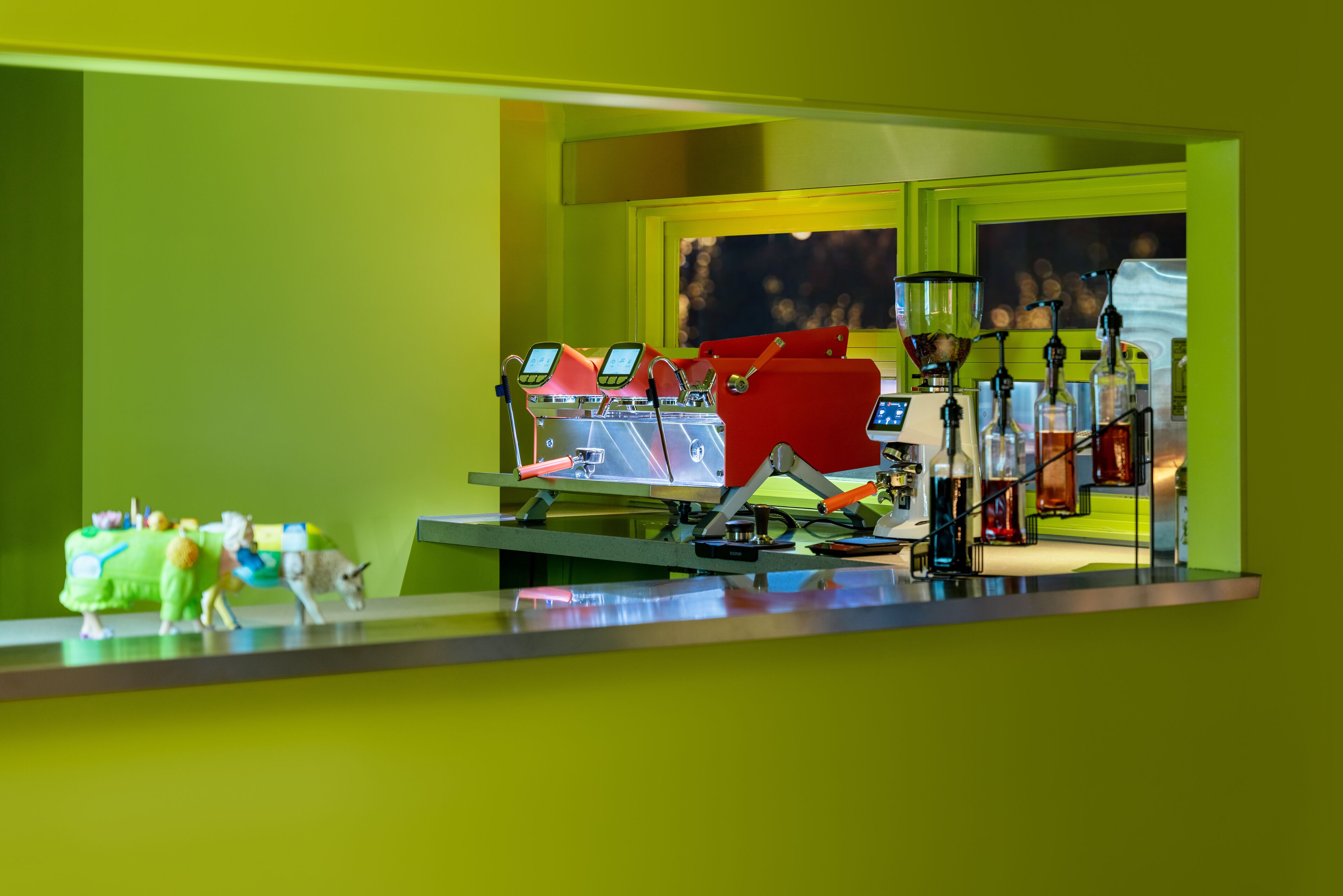 A bar area with multiple red bar stools, a counter with a digital screen, and various bottles and glasses.