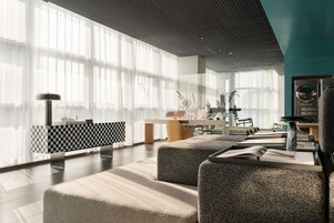 A modern hotel lobby with a checkered reception desk, seating area with ottomans, and a large window with sheer curtains.
