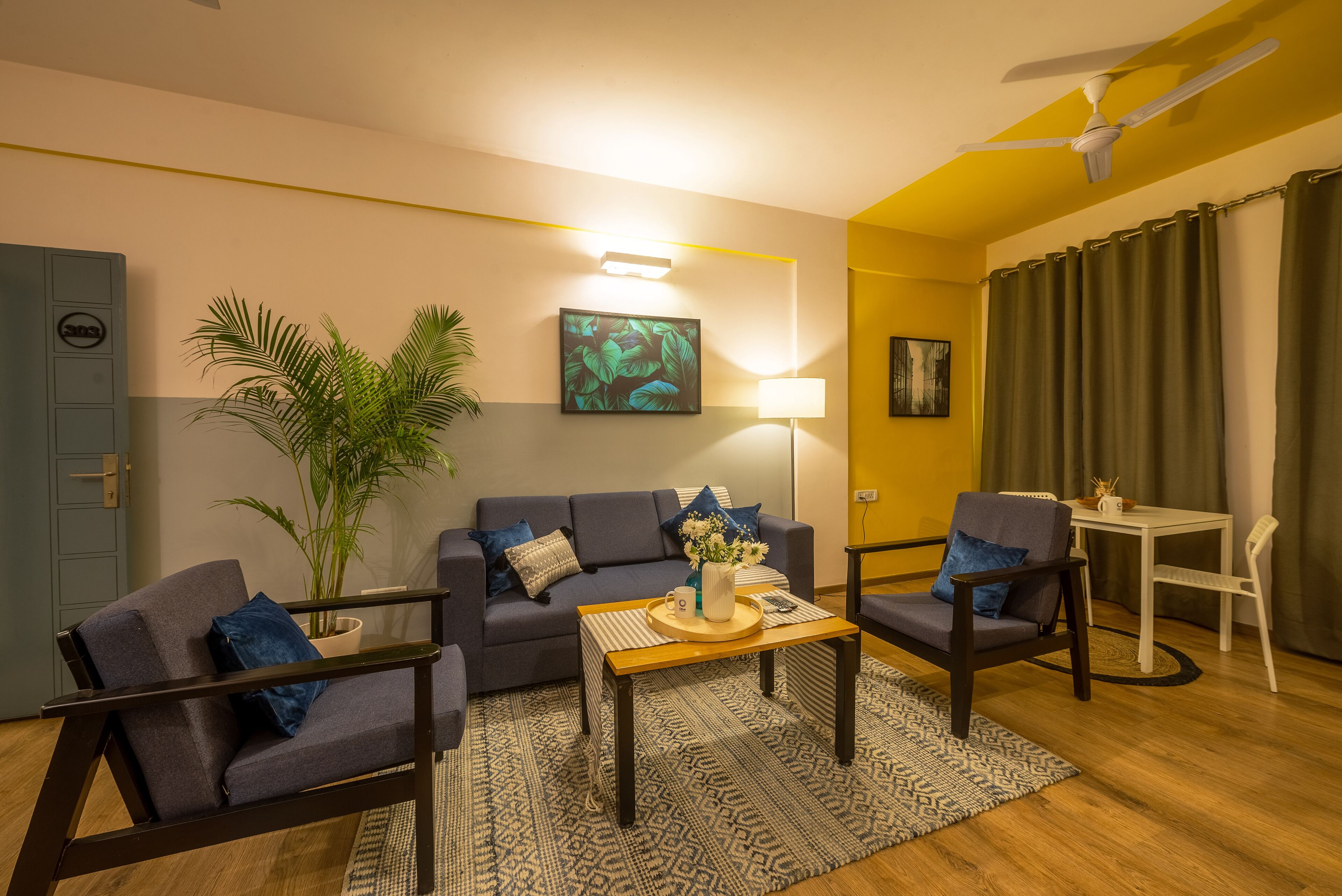 A modern living room with a sofa, two armchairs, a coffee table, a lamp, a plant, and a ceiling fan.