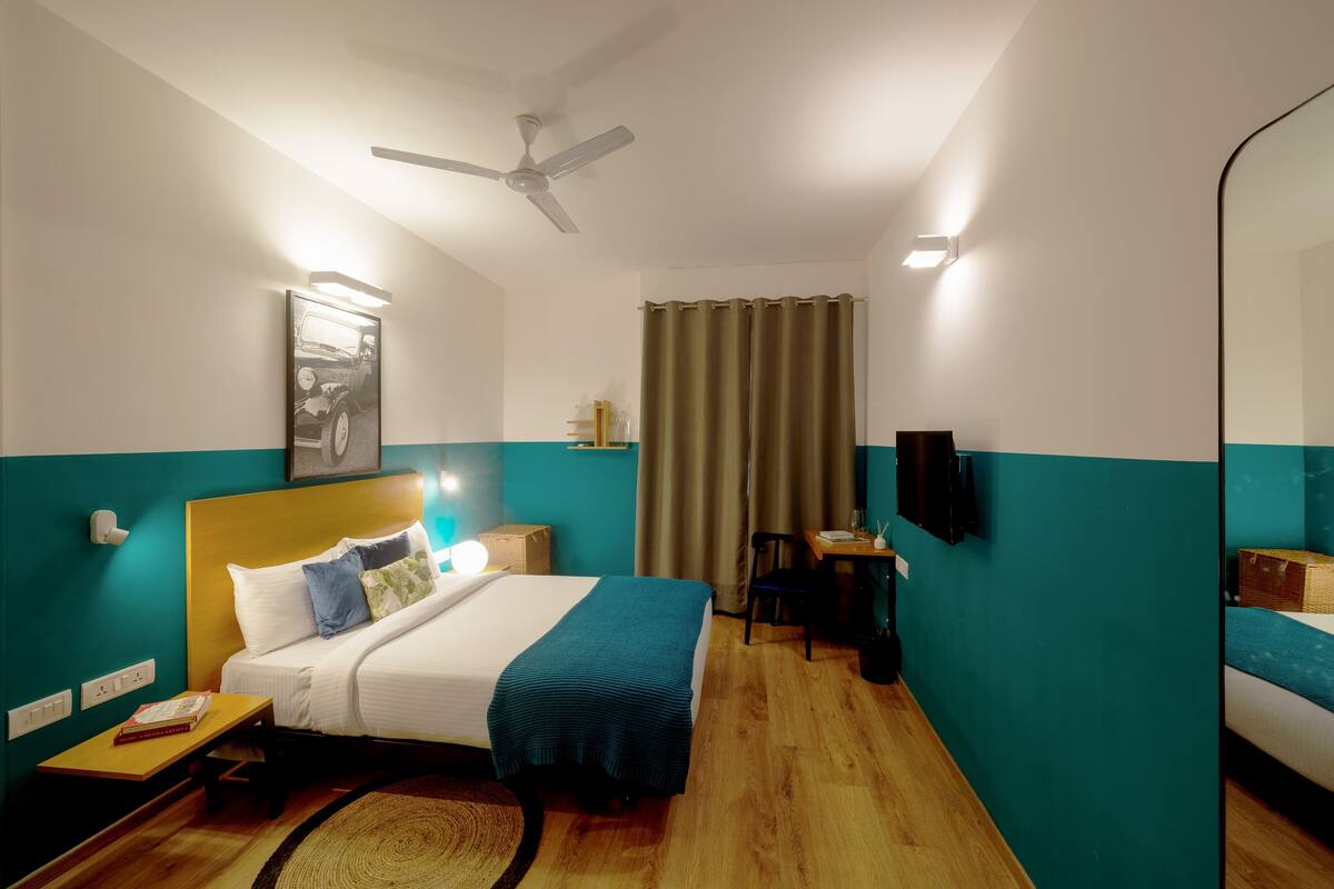 Habitación de hotel con cama, escritorio, silla, ventilador de techo, ventana con cortinas y televisor montado en la pared.