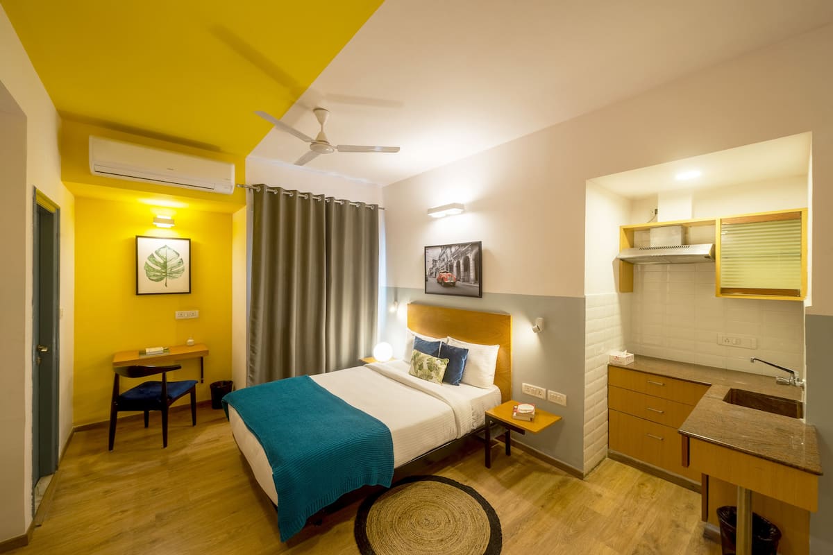 Habitación de hotel con cama, escritorio, silla, mesita, alfombra redonda, ventilador de techo, ventana con cortinas y kitchenette.