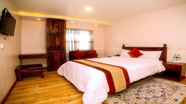 A bedroom with a wooden bed, a TV mounted on the wall, a wooden bench, and a patterned rug. - MeroStay 141 Hotel RG (Kathmandu)